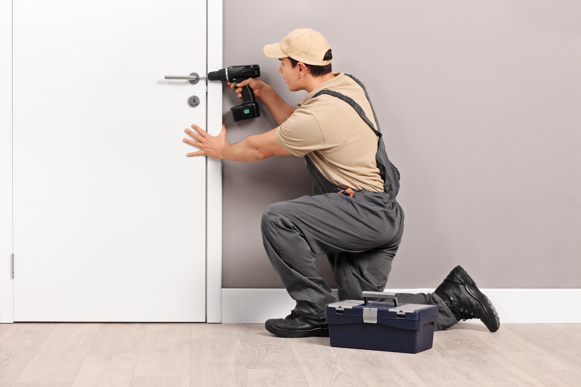 A technician installs a door handle using a cordless drill, crouching beside a toolbox on a wooden floor against a neutral wall.